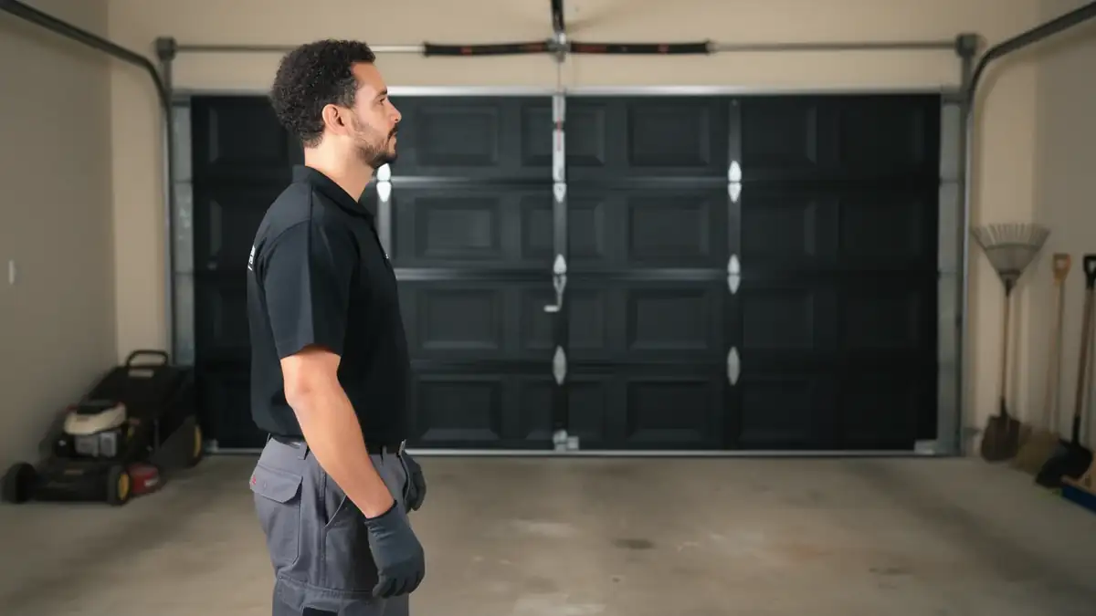 A technician stands in a garage with a closed black garage door behind him.